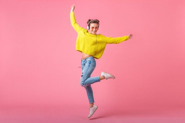 Jolie femme drôle heureuse sautant écouter de la musique dans des écouteurs habillés en tenue de style coloré hipster isolé sur un mur rose, portant un pull jaune et des lunettes de soleil, s'amuser