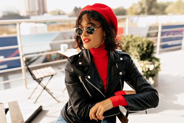 Photo gratuite jolie femme élégante en béret rouge et lunettes de soleil avec rouge à lèvres assis sur une terrasse au soleil