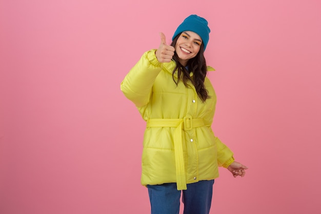 Jolie femme élégante excitée souriante montrant le pouce vers le haut posant en mode hiver look sur mur rose en veste jaune néon lumineux, portant un bonnet bleu, vêtu de vêtements chauds