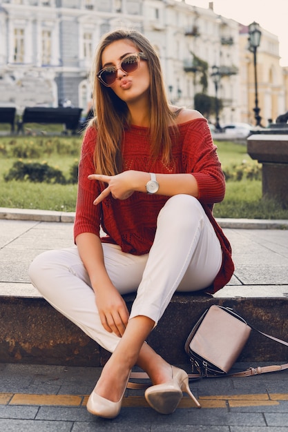 Jolie femme élégante en lunettes de soleil à la mode et pull rouge assis dans la rue