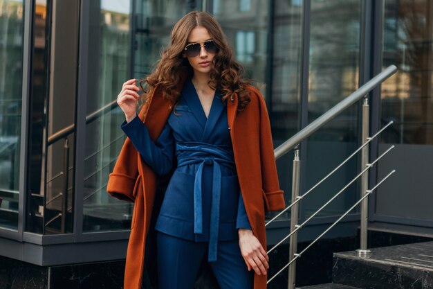 Jolie femme élégante avec marche dans la rue des affaires de la ville urbaine vêtue d'un manteau brun chaud et d'un costume bleu, style de rue mode tendance printemps automne, lunettes de soleil