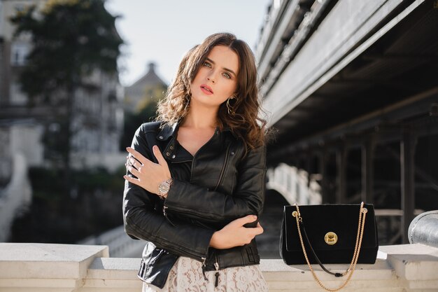 Jolie femme élégante posant dans la rue en tenue à la mode, sac à main en daim, vêtue d'une veste en cuir noir et robe en dentelle blanche, style printemps automne