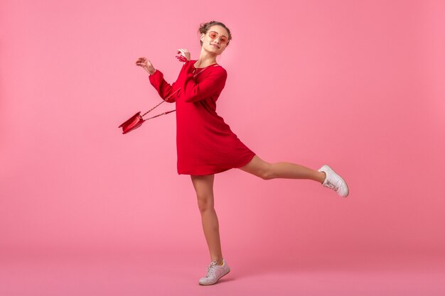 Jolie femme élégante souriante heureuse en robe à la mode rouge sautant en cours d'exécution sur un mur rose isolé, tendance de la mode printemps été, humeur romantique fille flirty