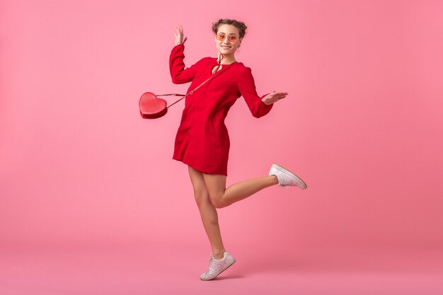 Jolie femme élégante souriante heureuse en robe à la mode rouge sautant en cours d'exécution sur un mur rose isolé, tendance de la mode printemps été, humeur romantique fille flirty