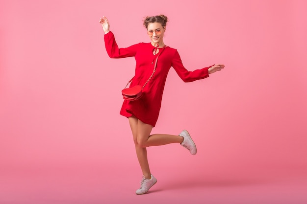 Jolie femme élégante souriante heureuse en robe à la mode rouge sautant en cours d'exécution sur un mur rose isolé, tendance de la mode printemps été, humeur romantique fille flirty