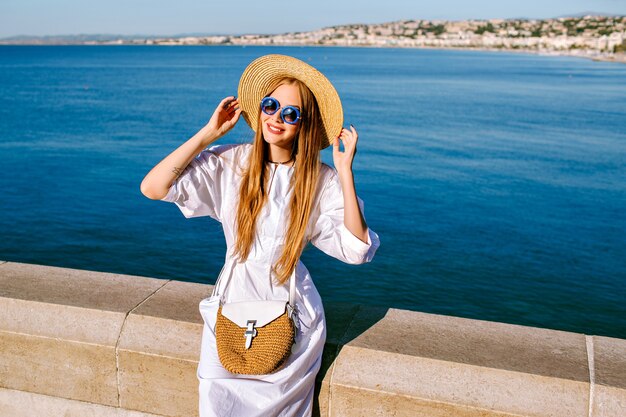 Jolie femme élégante vêtue d'une robe blanche, chapeau de paille et sac posant près de l'océan