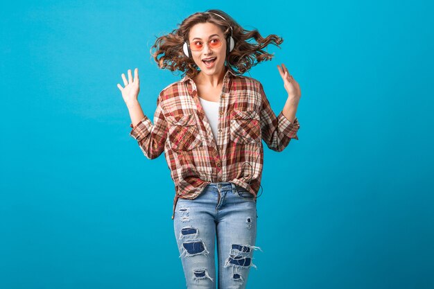 Jolie femme émotive souriante sautant avec l'expression du visage fou drôle en chemise à carreaux et jeans isolés sur fond bleu studio, portant des lunettes de soleil roses