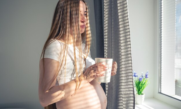 Une jolie femme enceinte avec une tasse de thé à la fenêtre le matin