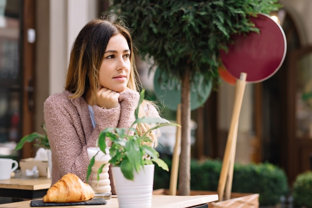 Jolie femme est assise dans la cafétéria vêtue d'un pull chaud et attend quelqu'un. Elle regarde de côté. Elle se réchauffe avec une boisson chaude. Journée d'automne, portrait à l'extérieur, rencontre.