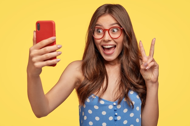 Jolie femme européenne heureuse aux cheveux longs, fait signe de paix, tient un téléphone portable, pose pour selfie, vêtue d'une robe bleue à pois, isolée sur un mur jaune fait un blog sur la vie quotidienne