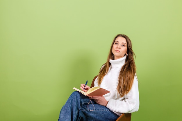 Jolie femme européenne en pull blanc décontracté isolé, mignon heureux s'asseoir sur une chaise avec bloc-notes et stylo