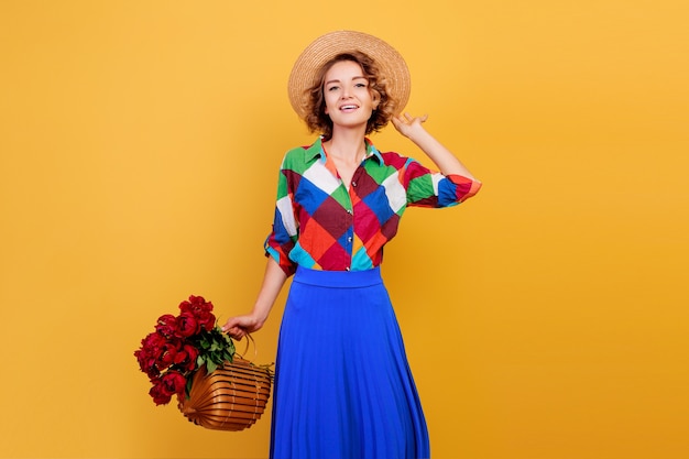 Jolie femme européenne en robe bleue tenant un bouquet de fleurs sur fond jaune. Chapeau de paille. Ambiance d'été.