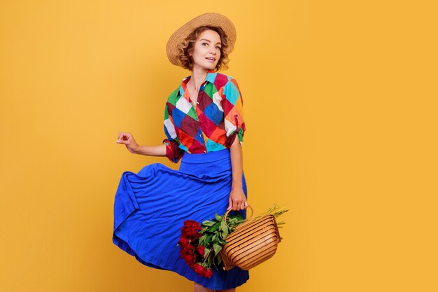 Jolie femme européenne en robe bleue tenant un bouquet de fleurs sur fond jaune. Chapeau de paille. Ambiance d'été.