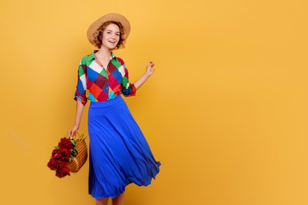 Jolie femme européenne en robe bleue tenant un bouquet de fleurs sur fond jaune. Chapeau de paille. Ambiance d'été.