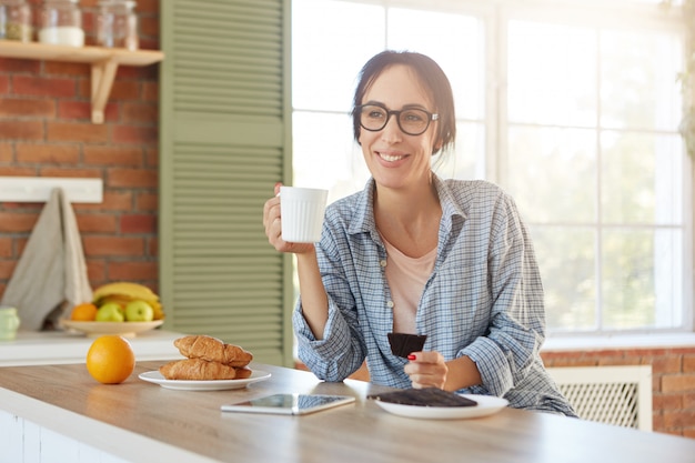 Jolie femme a une expression heureuse aime le café du matin avec de délicieux croissants sucrés et du chocolat