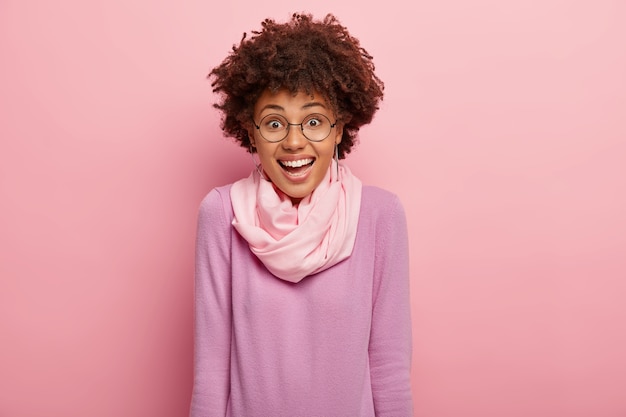 Jolie femme frisée exprime la joie, profite d'un moment drôle, rit de quelque chose, sourit positivement, porte des lunettes rondes