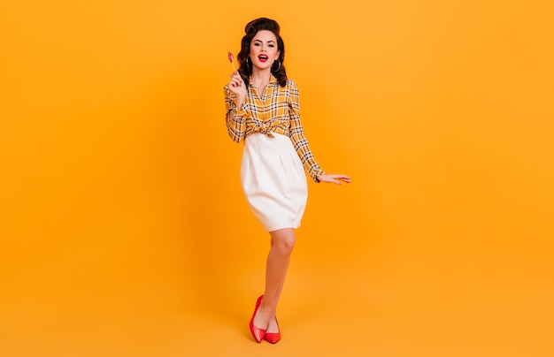 Jolie femme galbée en jupe blanche posant sur fond jaune. Photo de Studio de pin-up brune tenant des bonbons.