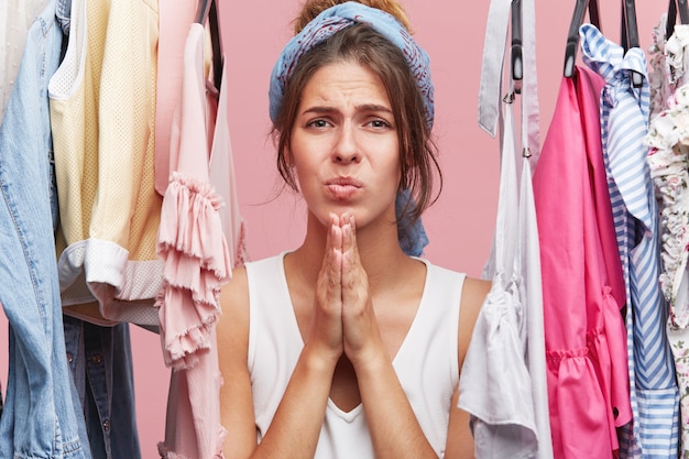 Jolie femme gardant les mains ensemble à la recherche avec une expression douloureuse, debout près de sa garde-robe, se plaignant qu'elle n'a pas de robes