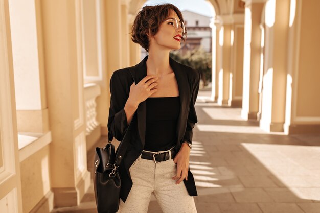 Jolie femme en haut noir, veste et pantalon blanc avec ceinture posant à l'extérieur. Femme aux cheveux ondulés avec sac à main et lunettes détourne le regard en ville.