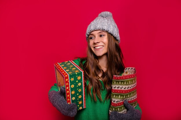 Jolie femme heureuse avec de longs cheveux brun clair et un sourire merveilleux habillé bonnet gris d'hiver, mitaines et pull vert tenant des cadeaux de nouvel an et souriant sur fond rouge isolé