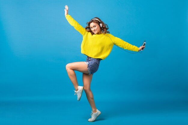 Jolie femme heureuse souriante sautant danser en écoutant de la musique dans des écouteurs en tenue de hipster isolé sur fond bleu studio, portant des shorts et un pull jaune