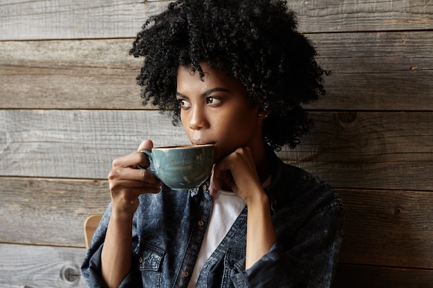 Photo gratuite jolie femme hipster afro-américaine habillée avec élégance, buvant du café ou du thé pensivement dans une grande tasse, détournant les yeux avec une expression pensive sérieuse, faisant des plans pour la journée les gens et le style de vie