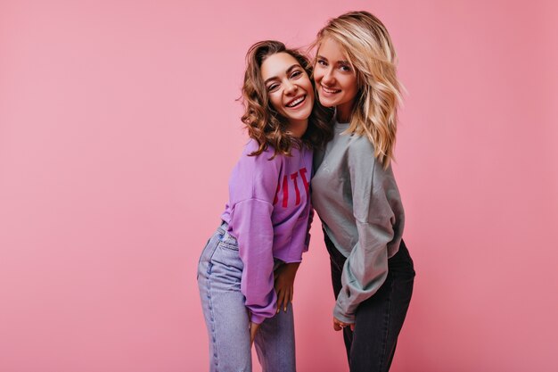 Jolie femme en jeans vintage riant avec sa soeur. Portrait intérieur de filles blithesome debout sur rose avec sourire.