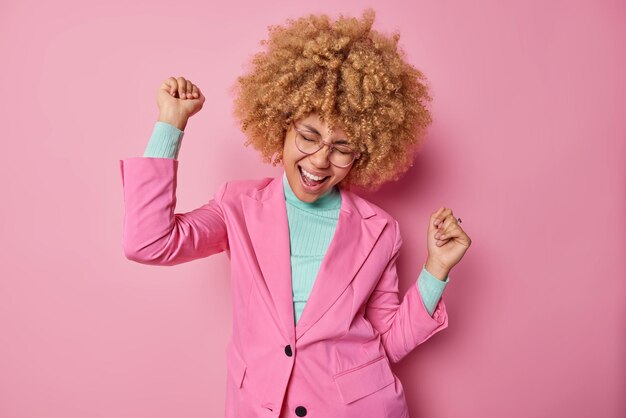 Une jolie femme joyeuse aux cheveux bouclés donne l'impression que la danse du triomphe est vêtue de vêtements élégants célèbre ses réalisations personnelles isolées sur fond rose Enfin, j'ai atteint mon objectif