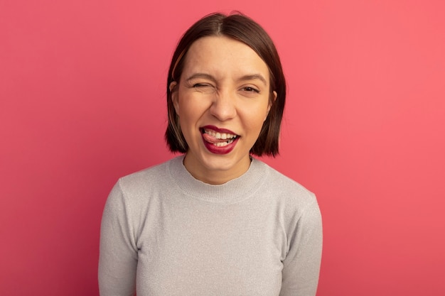 Jolie femme joyeuse clignote des yeux et sort la langue isolée sur le mur rose