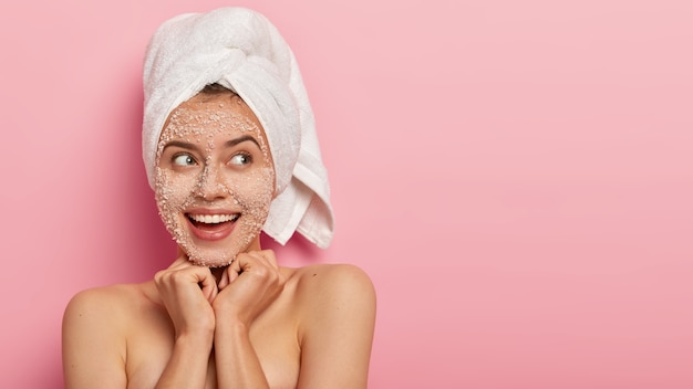 Jolie femme joyeuse garde les deux mains sous le menton, a une expression rêveuse, porte une serviette blanche douce sur la tête, a un corps nu