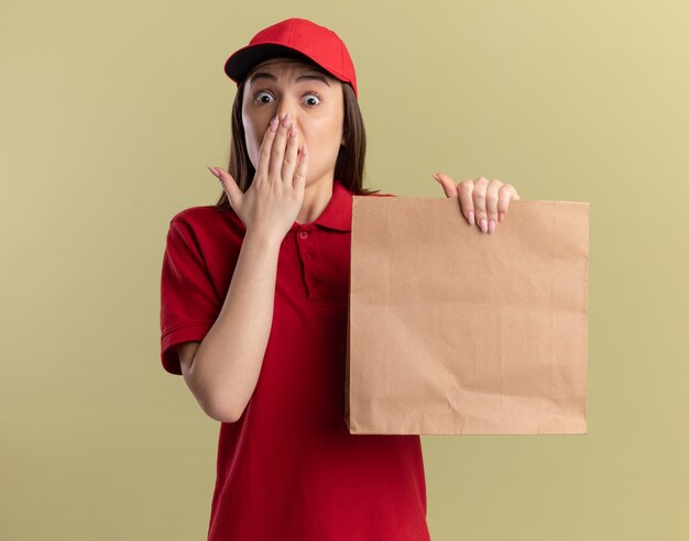 Jolie femme de livraison anxieuse en uniforme met la main sur la bouche et détient un paquet de papier sur vert olive