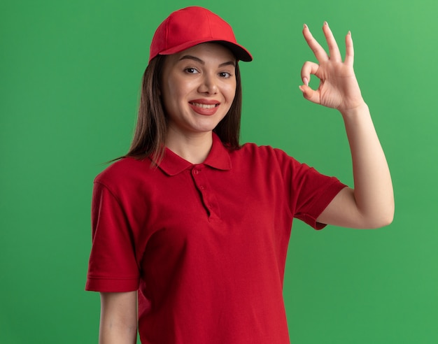 Jolie femme de livraison souriante en gestes uniformes signe de main ok isolé sur mur vert avec espace de copie