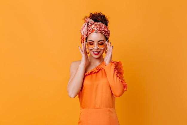 Jolie femme à lunettes orange, robe en soie et bandeau sourit sur l'espace orange.