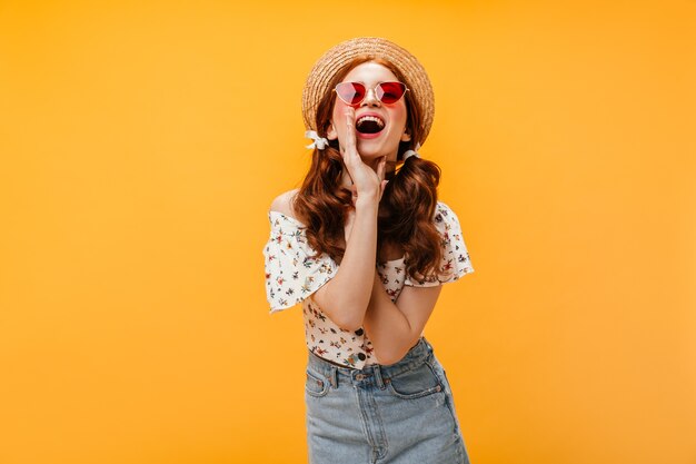 Jolie femme en lunettes de soleil rouges et chapeau hurle. Dame habillée en jupe en jean, t-shirt blanc et chapeau posant sur fond orange.