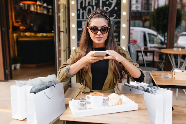 Jolie femme en lunettes de soleil sombres assis dans un café en plein air à côté de sacs de boutique