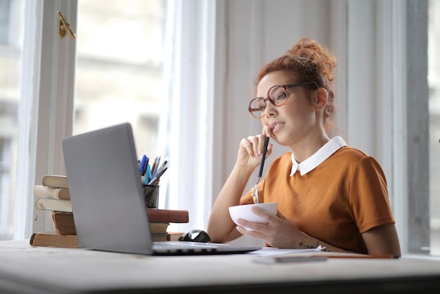 Jolie Femme Avec Des Lunettes Tenant Un Bol De Céréales Et Assis Devant Un Ordinateur Portable Sur Le Bureau