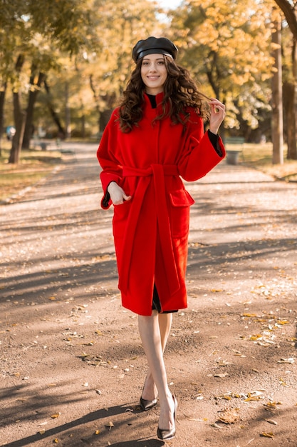 Jolie femme maigre souriante élégante avec des cheveux bouclés marchant dans le parc habillé en manteau rouge chaud automne mode tendance, style de rue, portant un béret