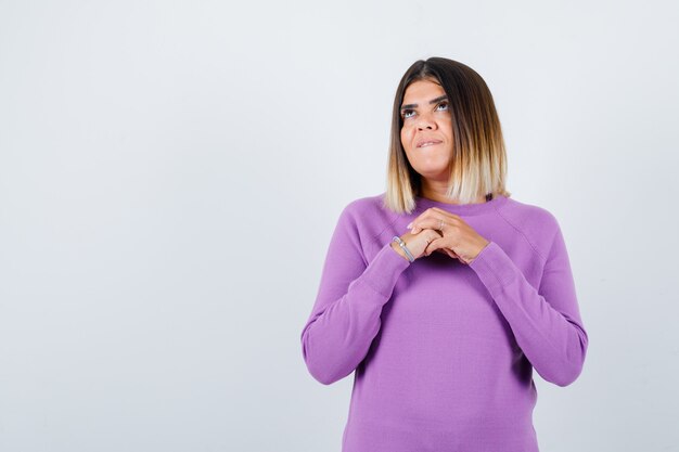 Jolie femme avec les mains sur la poitrine, levant les yeux, mordant la lèvre en pull violet et l'air rêveuse. vue de face.