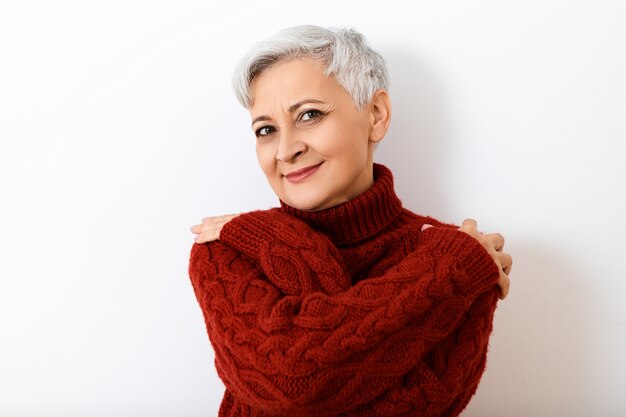 Jolie femme mature senior aux cheveux courts avec birhgt maquillage posant isolé portant élégant pull en tricot, se serrant dans ses bras, ayant une expression faciale joyeuse et joyeuse