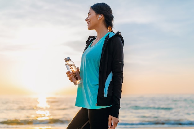 Jolie femme mince faisant des exercices de sport sur la plage du lever du soleil du matin en vêtements de sport, eau potable assoiffée en bouteille, mode de vie sain, écouter de la musique sur des écouteurs sans fil, sourire heureux