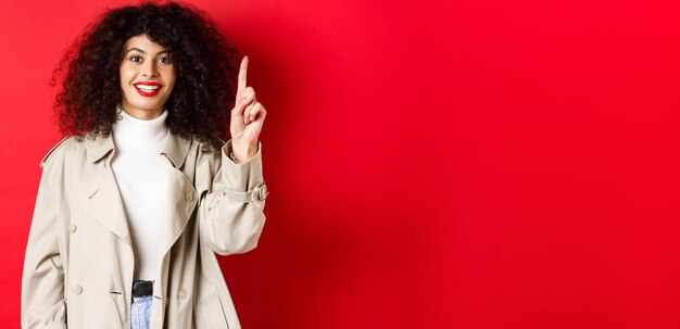 Jolie femme moderne avec des lèvres rouges coiffure frisée portant un trench-coat de printemps pointant le doigt vers le haut