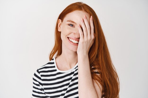 Jolie femme moel aux cheveux roux et aux yeux bleus souriant, couvrant la moitié du visage avec la main, debout sur un mur blanc