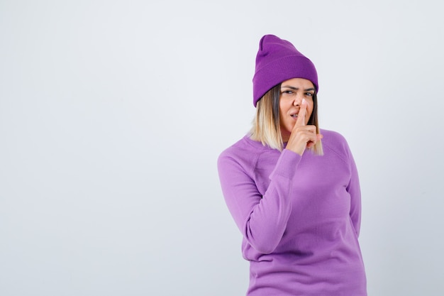 Jolie femme montrant un geste de silence en pull, bonnet et semblant sensible, vue de face.