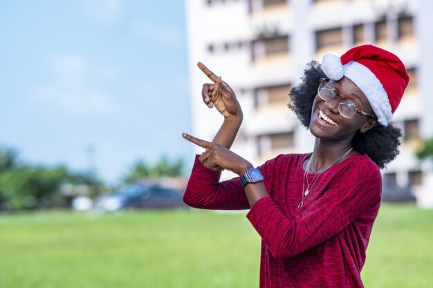 Jolie femme noire séduisante souriante dans un bonnet de Noel et des lunettes pointant ses doigts vers le ciel