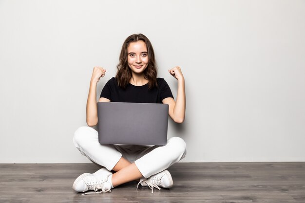 Jolie femme avec un ordinateur portable moderne assis sur le sol avec un geste de victoire sur un mur gris