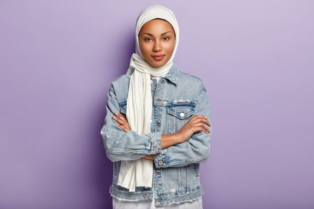 Une jolie femme orientale couvre la tête avec un foulard blanc pour garder sa dignité et son pouvoir, a un code vestimentaire spécial, garde les mains croisées, regarde avec modestie, pose sur un mur violet. Règles islamiques