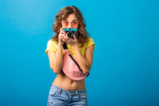Jolie femme posant avec appareil photo vintage à prendre des photos habillées en tenue colorée d'été hipster isolée sur fond bleu, cachant l'expression du visage surpris