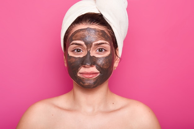 Une jolie femme pose avec une expression faciale sérieuse et calme, a un masque hocolate sur le visage, les épaules nues, prend soin de sa beauté et de son apparence, porte une serviette blanche sur la tête. Concept de soins de la peau.