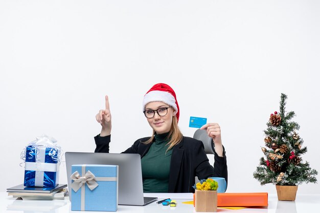 Jolie femme positive avec chapeau de père Noël et portant des lunettes assis à une table cadeau de Noël et tenant une carte bancaire pointant au-dessus dans le bureau