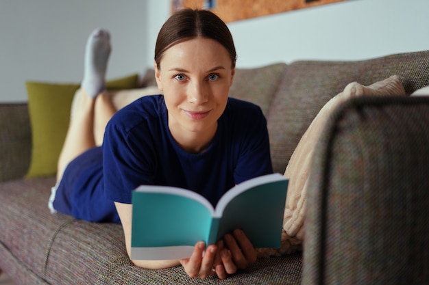 Jolie femme positive lisant un livre de détente sur le canapé